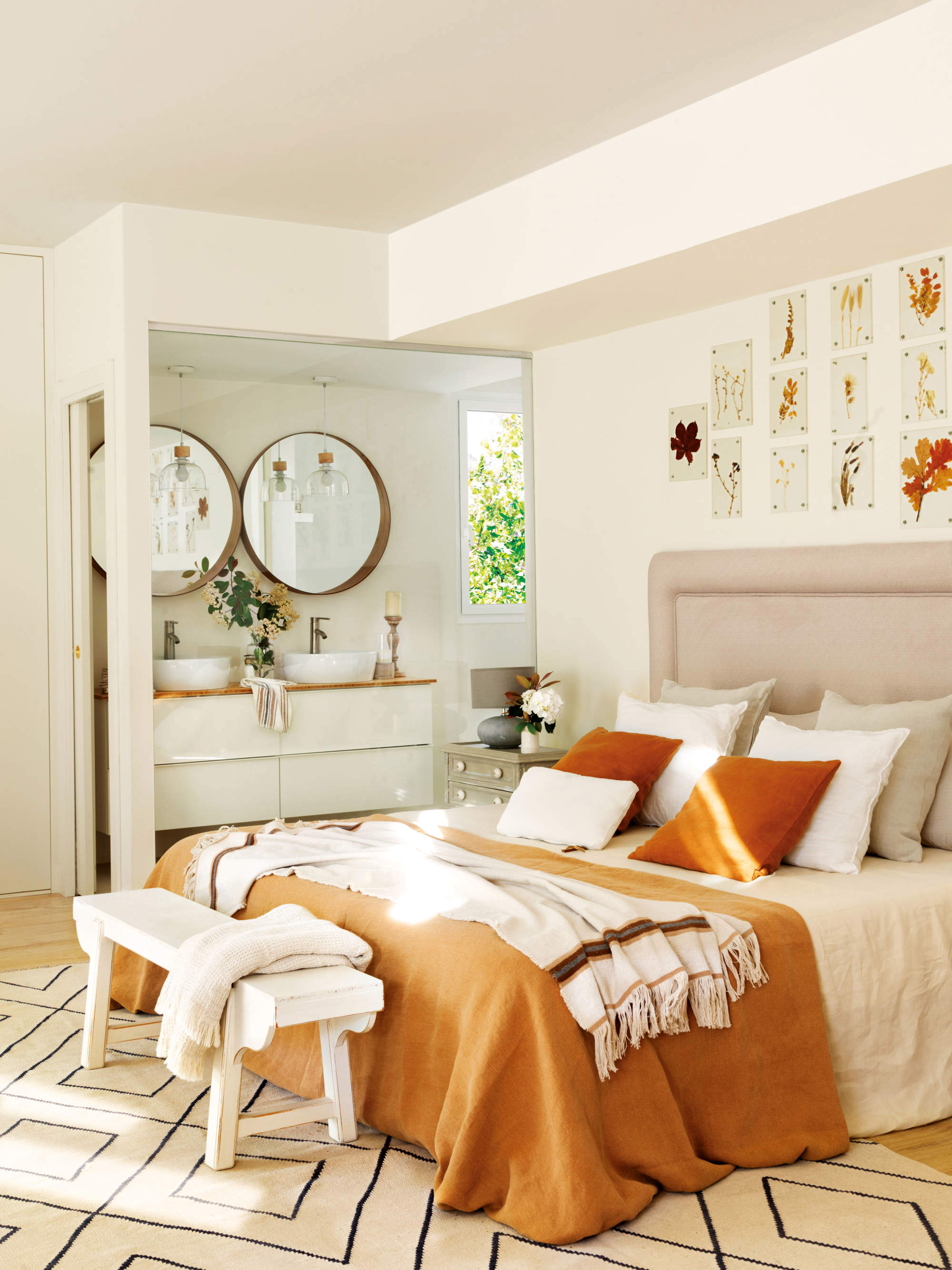 Dormitorio blanco con ropa de cama en tonos naranjas, baño con tabique acristalado, y decoración floral seca.