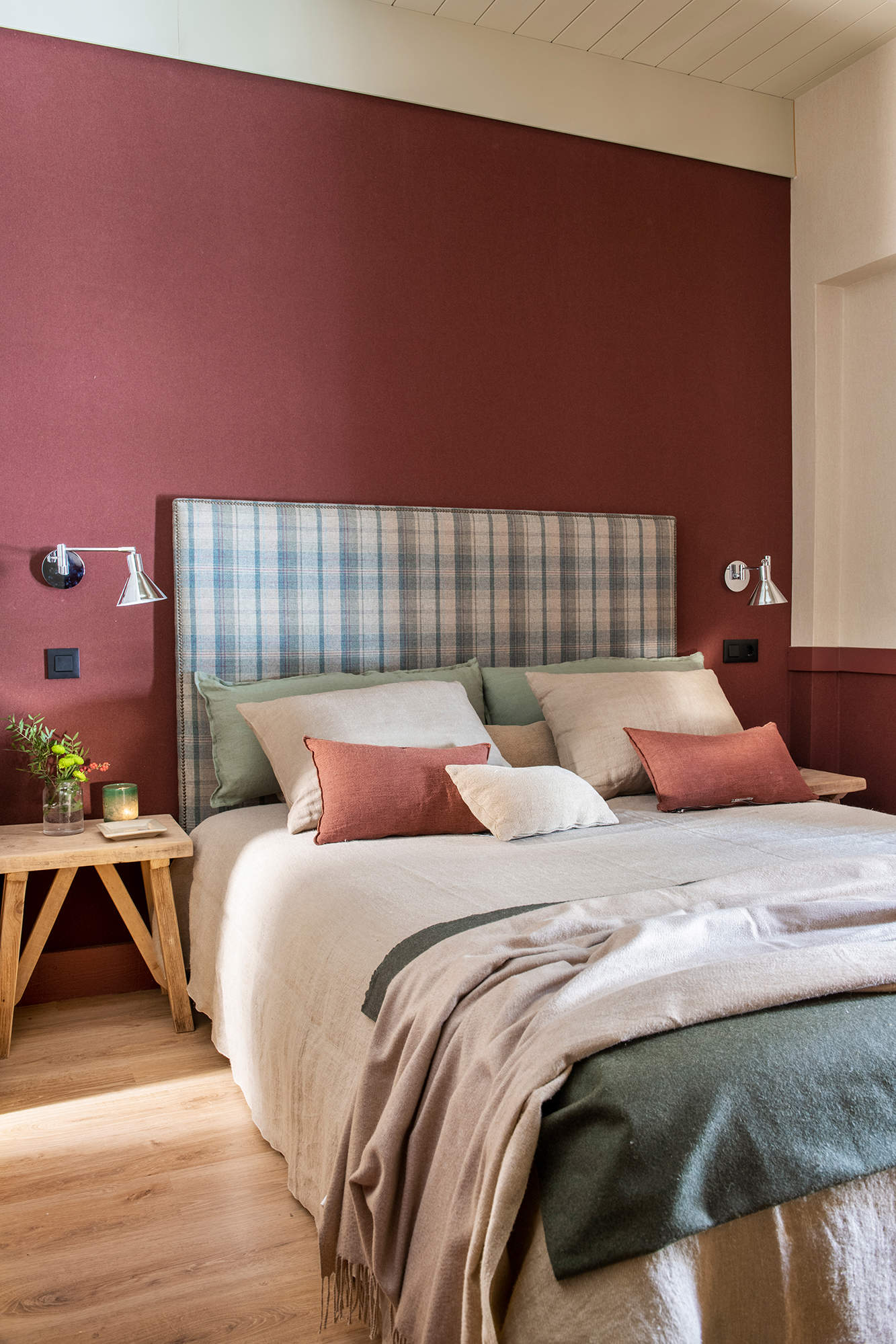Dormitorio con pared del cabecero pintado en granate, cabecero tapizado de cuadros, mesilla de madera y ropa de cama en beige y verde.