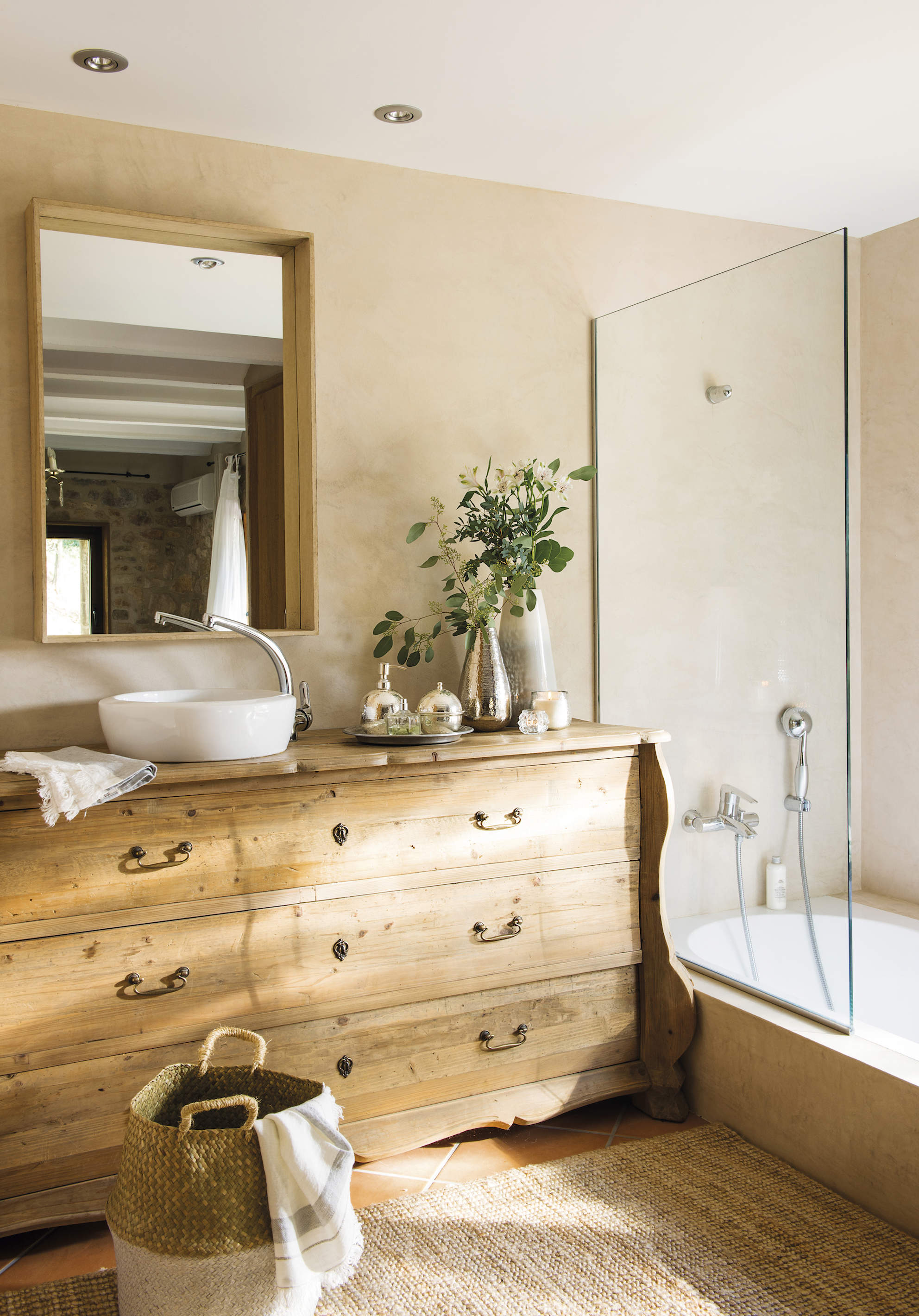 Baño con cómoda de madera como mueble bajolavabo, ducha con mampara transparente, bañera, lavabo sobreencimera, espejo, cesta y alfombra de fibra 