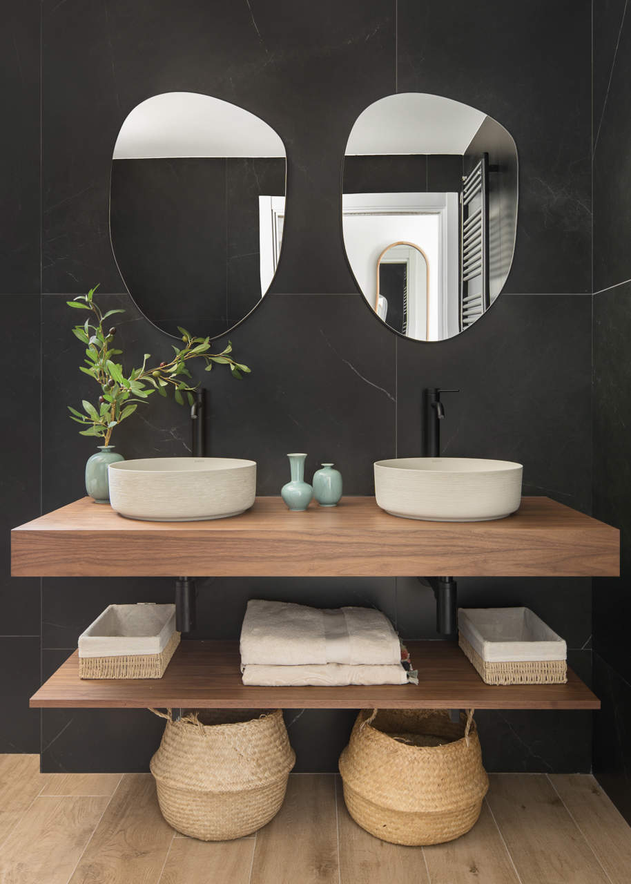 Baño doble con baldas de madera, paredes negras y espejos desiguales.