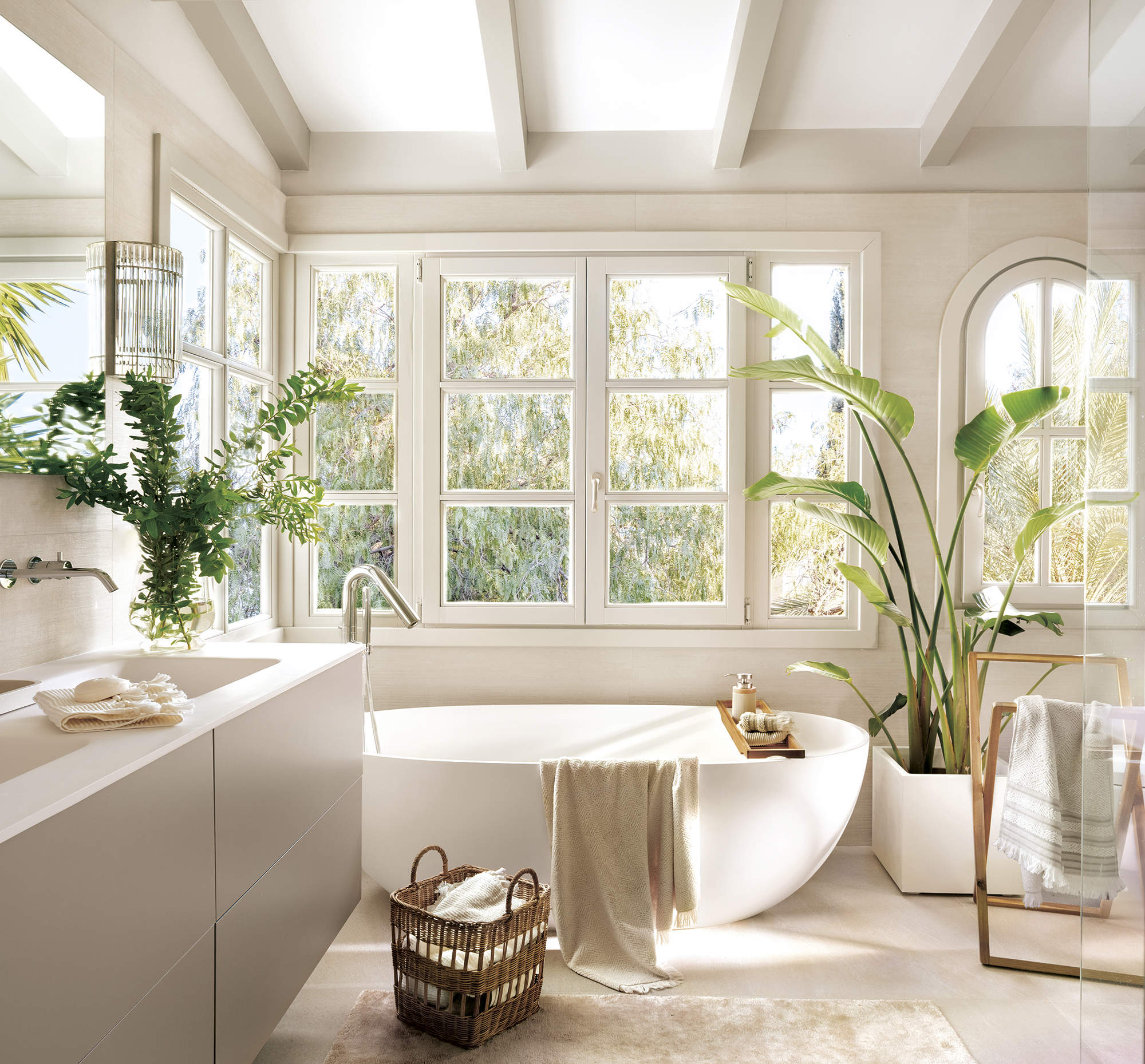 baño grande con bañera externa blanca y mueble de lavabo de dos senos, plantas, suelo de piedra con alfombra beige, gran ventanal.