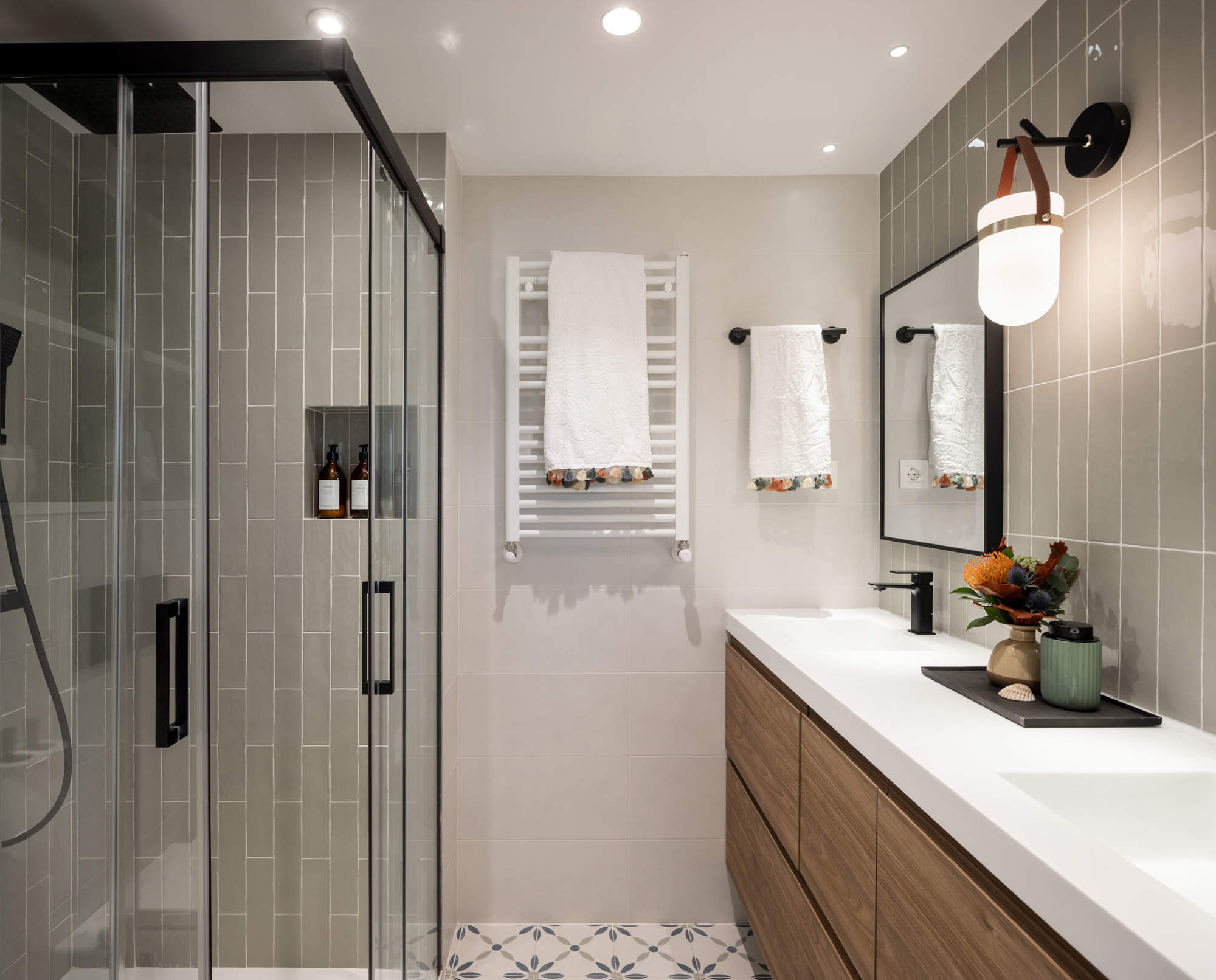 Baño con ducha con perfilería negra, mueble de madera, azulejos con flores en el suelo y azulejos verdosos en la pared.
