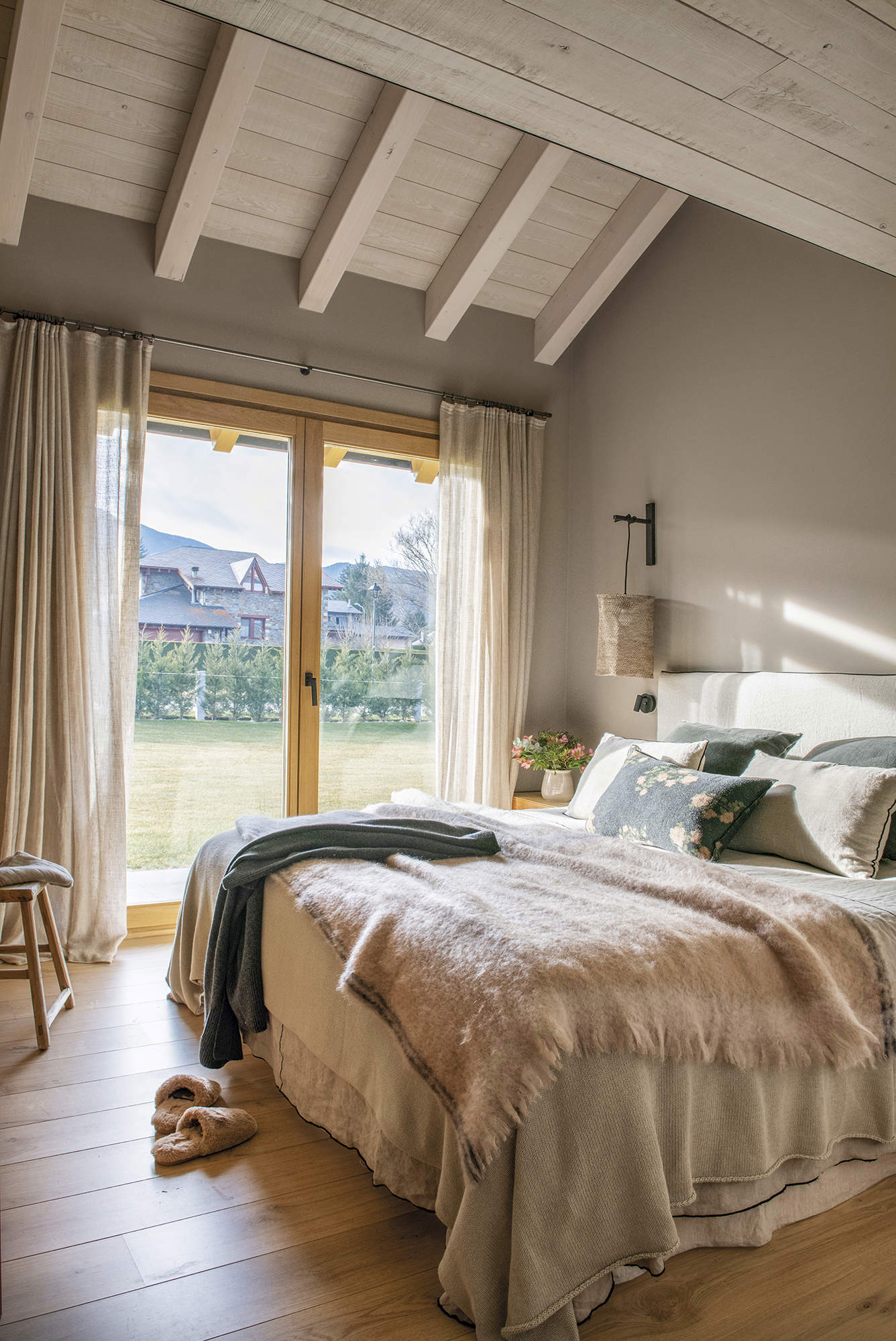 Dormitorio rústico con ropa de cama en rosa palo y techo abuhardillado con vigas de madera. 