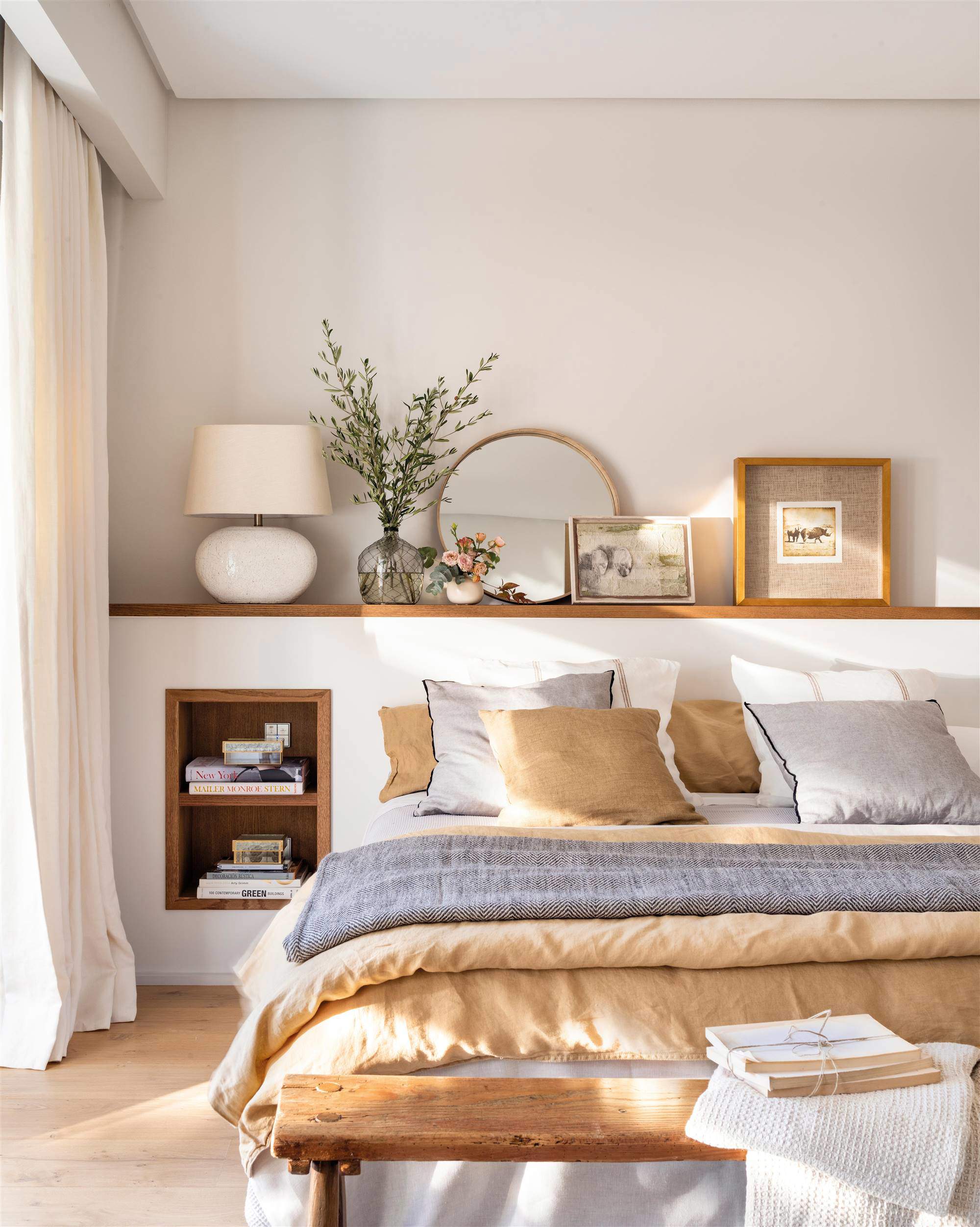 Dormitorio con cabecero de obra y una composición de cuadros, flores y un espejo redondo en la repisa del cabecero.