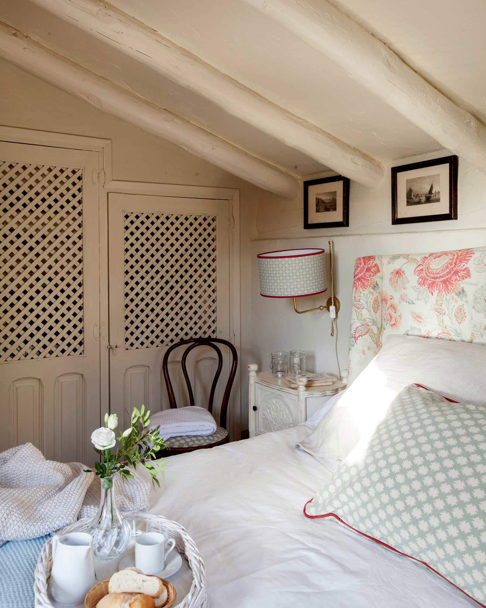 Dormitorio abuhardillado con cabecero tapizado y estampado floral, armarios empotrados con puerta de rejilla y aplique.