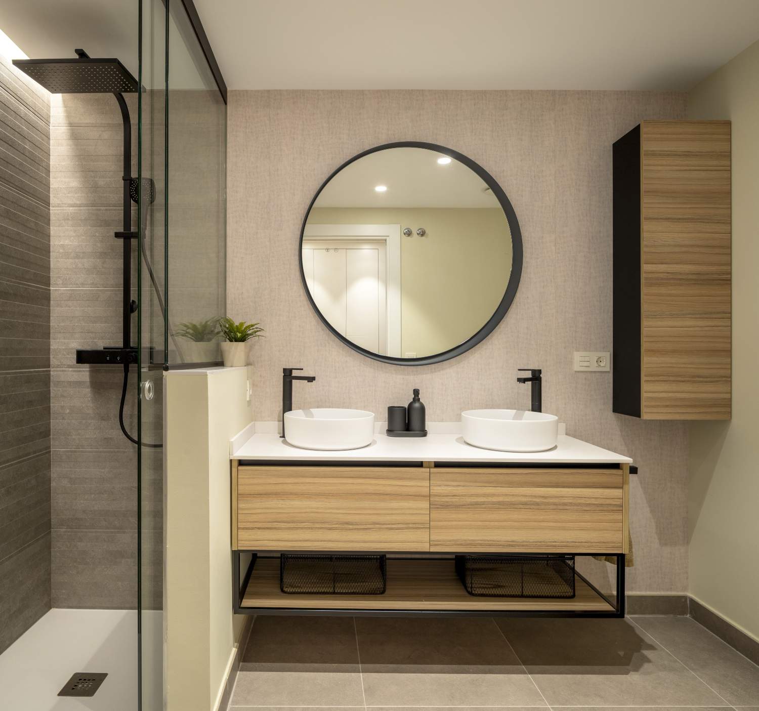 Baño moderno con espejo redondo y ducha con mampara con detalles negros.