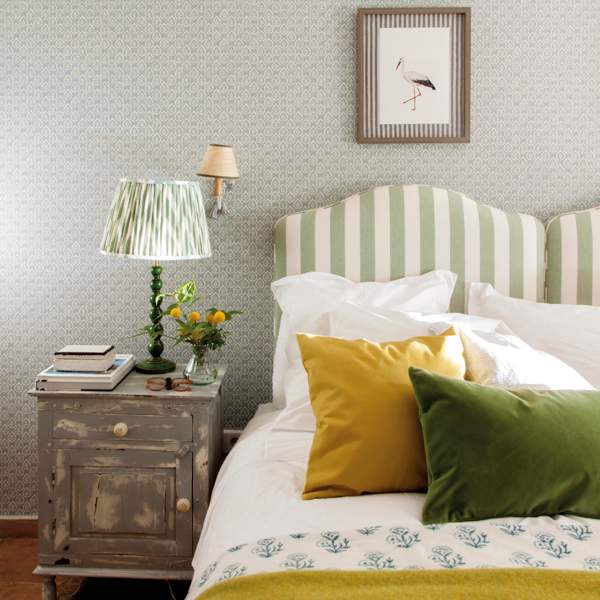 Dormitorio con cabecero a rayas verdes, lámpara de sobremesa plisada, mesilla de noche recuperada y cuadro 