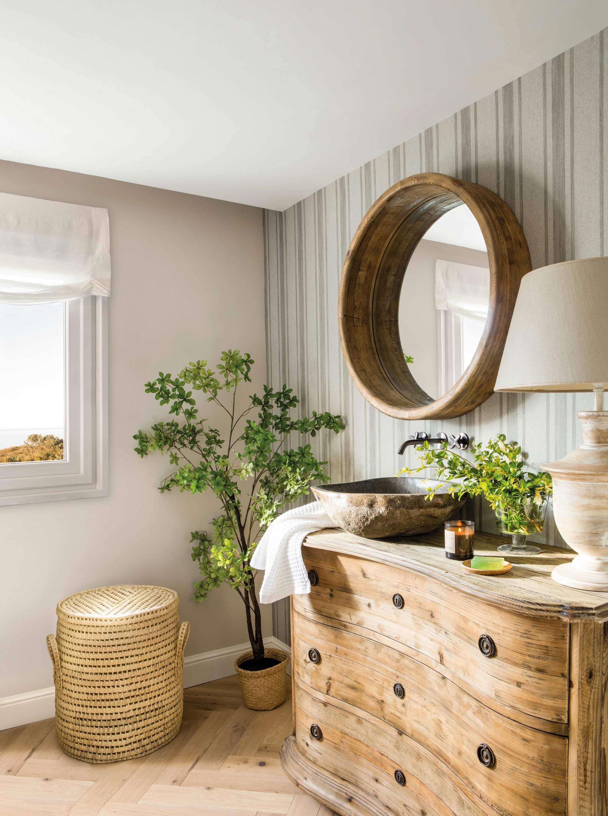 Baño con cómoda ondulada de madera como mueble de lavabo, lavamanos exento, espejo redondo, cesto de ropa, plantas y lámpara de mesa