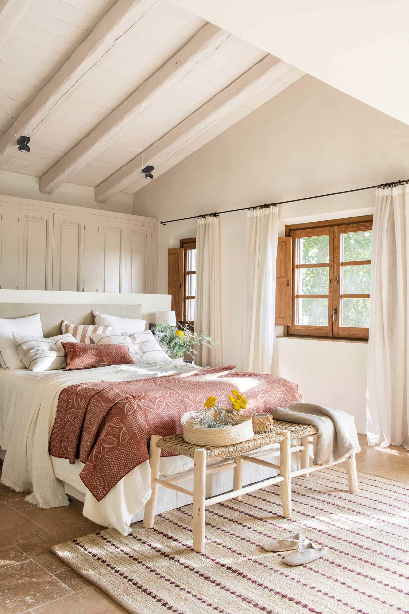 Dormitorio pequeño abuhardillado con ventanas de madera, banco a los pies y alfombra.