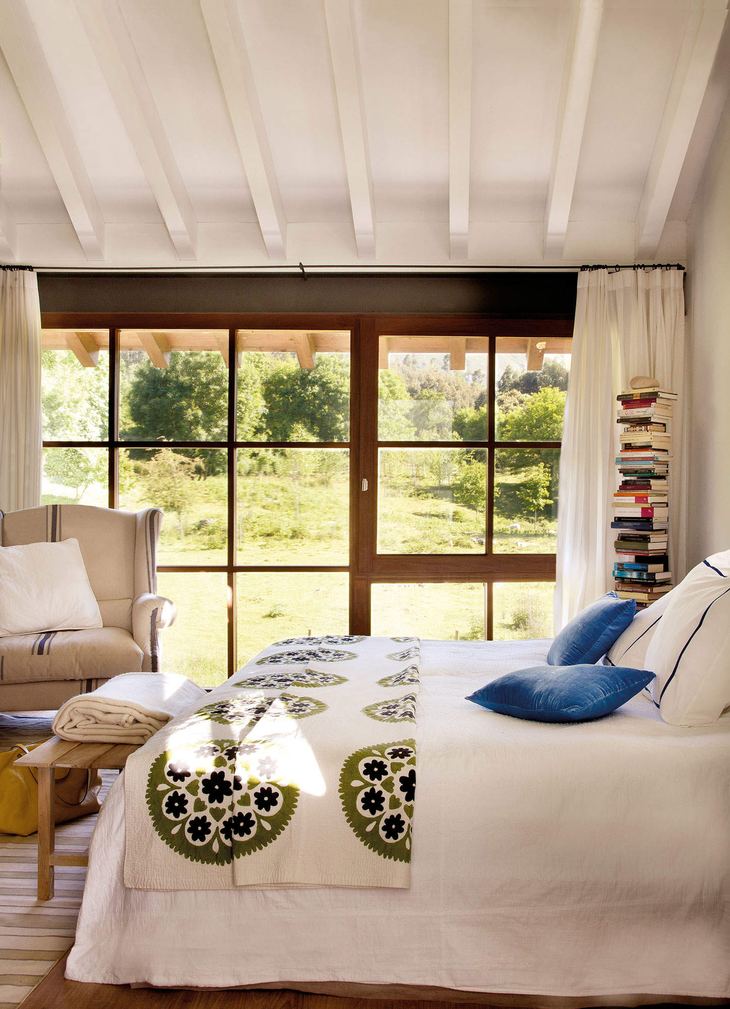 Dormitorio con vigas de madera en blanco y gran ventana al jardín con estantería tipo columna