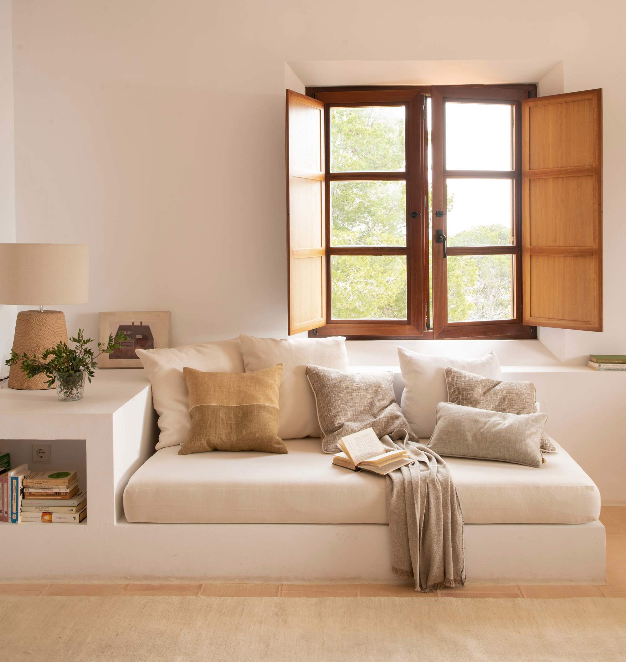 Salón con sofá de obra, colchoneta, cojines, plaid, ventana y lámpara de sobremesa
