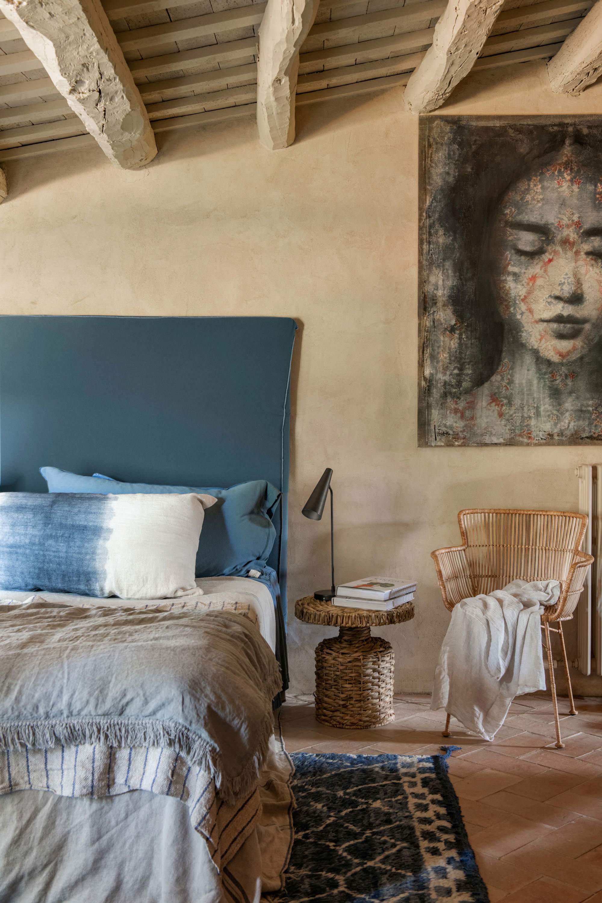 Dormitorio de inspiración asiática con vigas de madera, cabecero tapizado azul, cuadro, mesa y silla de fibra