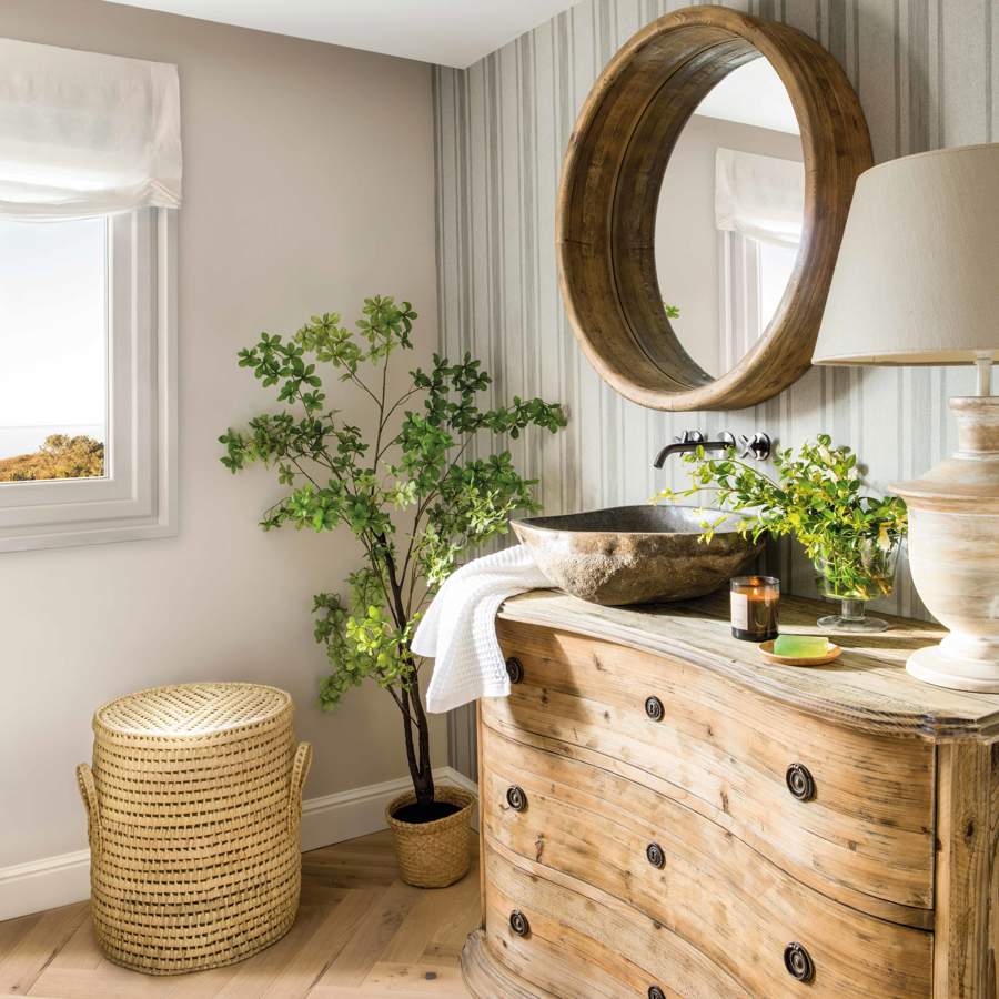 Baño con cómoda ondulada de madera como mueble de lavabo, lavamanos exento, espejo redondo, cesto de ropa, plantas y lámpara de mesa