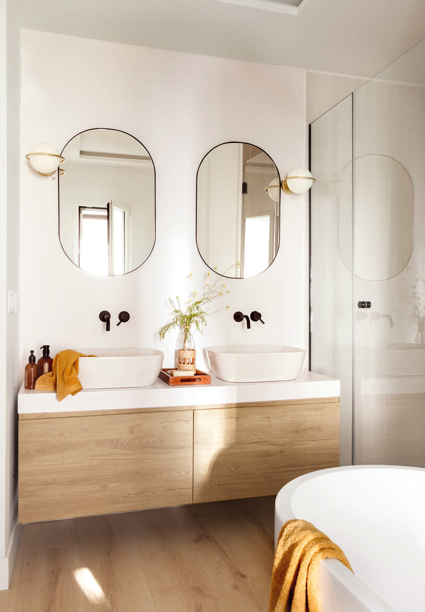 Baño pequeño con bañera, mueble volado, lavabos exentos, grifería empotrada, espejos ovalados y apliques globo.