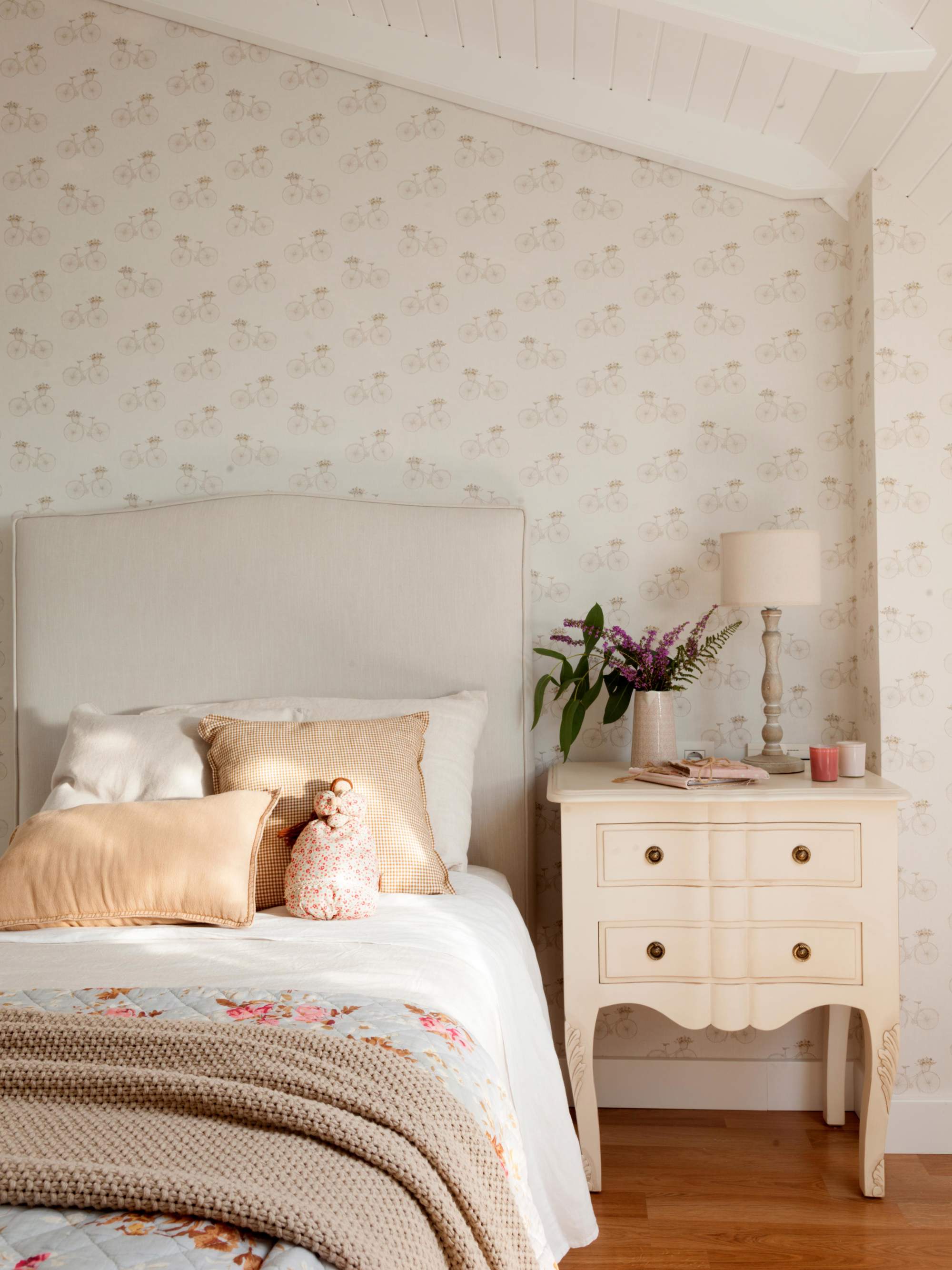 Dormitorio abuhardillado con papel pintado en cabecero y mesita de noche blanco con cajones