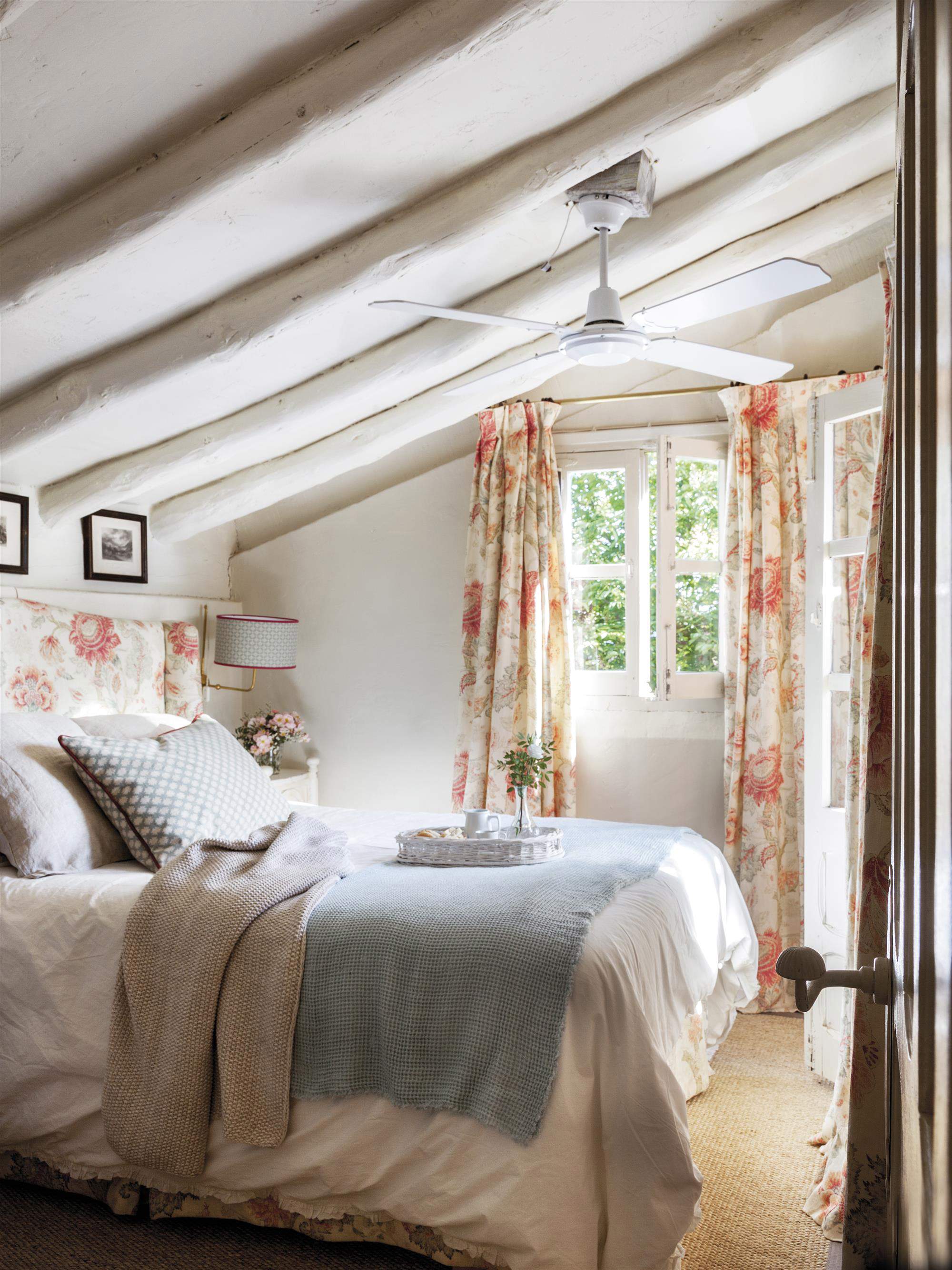 Dormitorio abuhardillado con vigas lacadas en blanco y cabecero y cortinas de flores