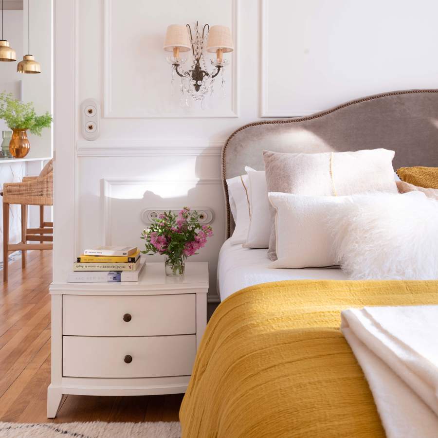Dormitorio con molduras, mesilla blanca, cabecero tapizado en gris con ribete, apliques antiguos y colcha mostaza             colcha mostaza 