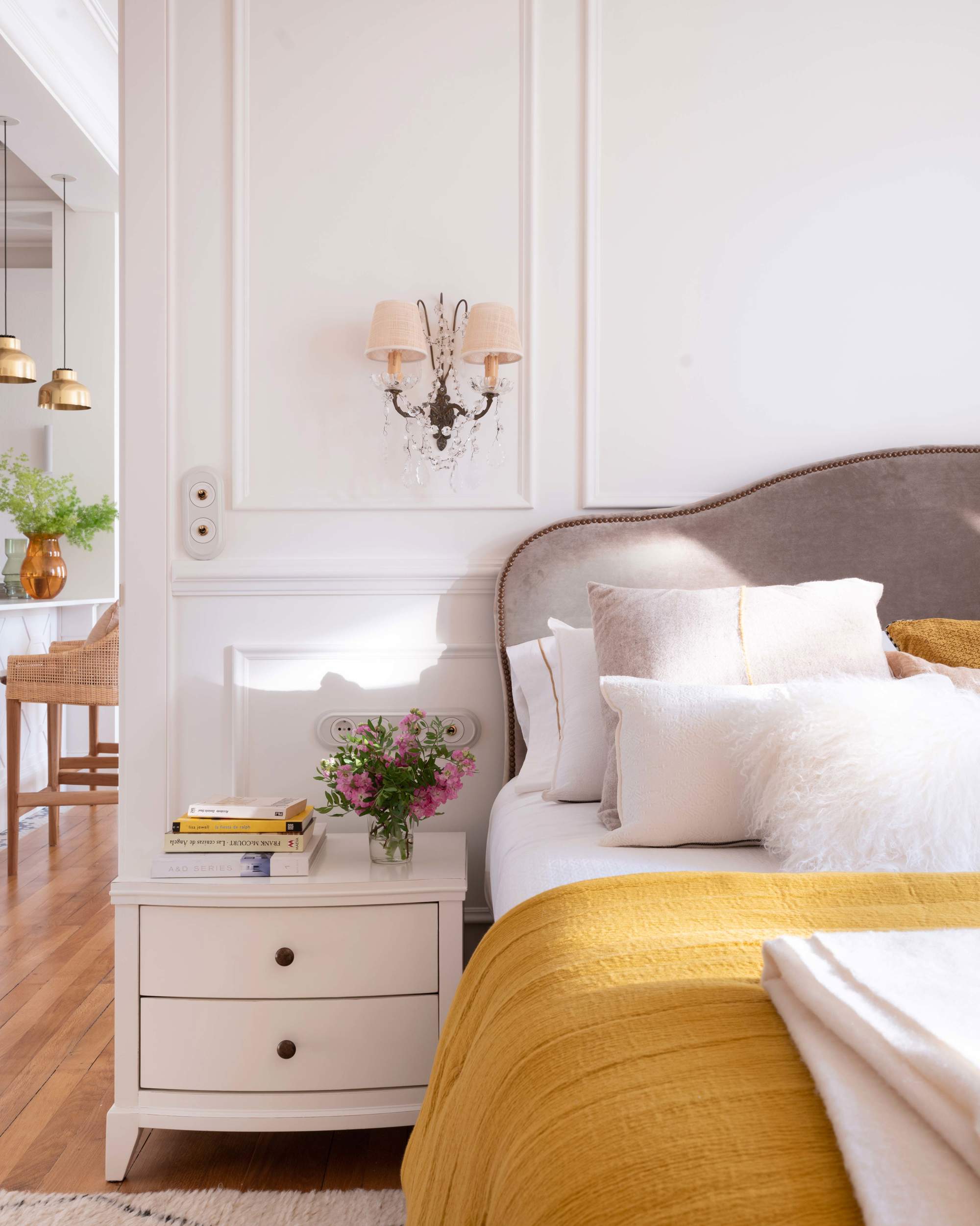 Dormitorio con molduras, mesilla blanca, cabecero tapizado en gris con ribete, apliques antiguos y colcha mostaza             colcha mostaza 