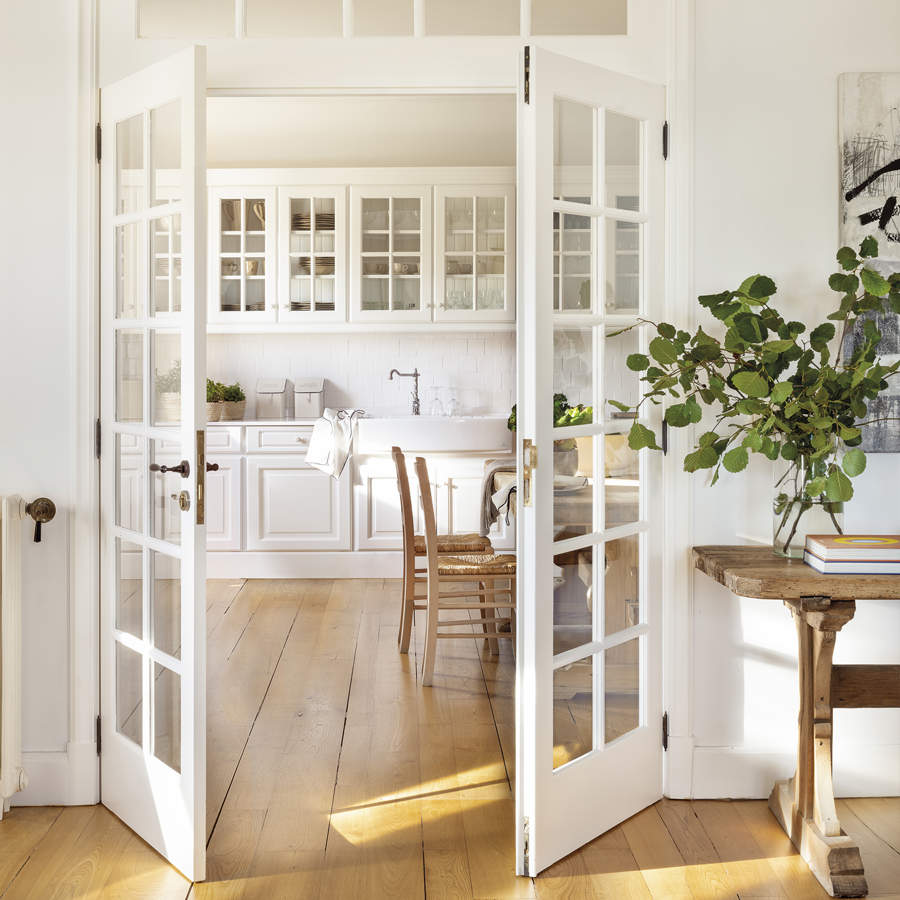 cocina con puertas acrisoladas blancas, suelo de madera natural con muebles blancos, mesa y sillas de madera, planta