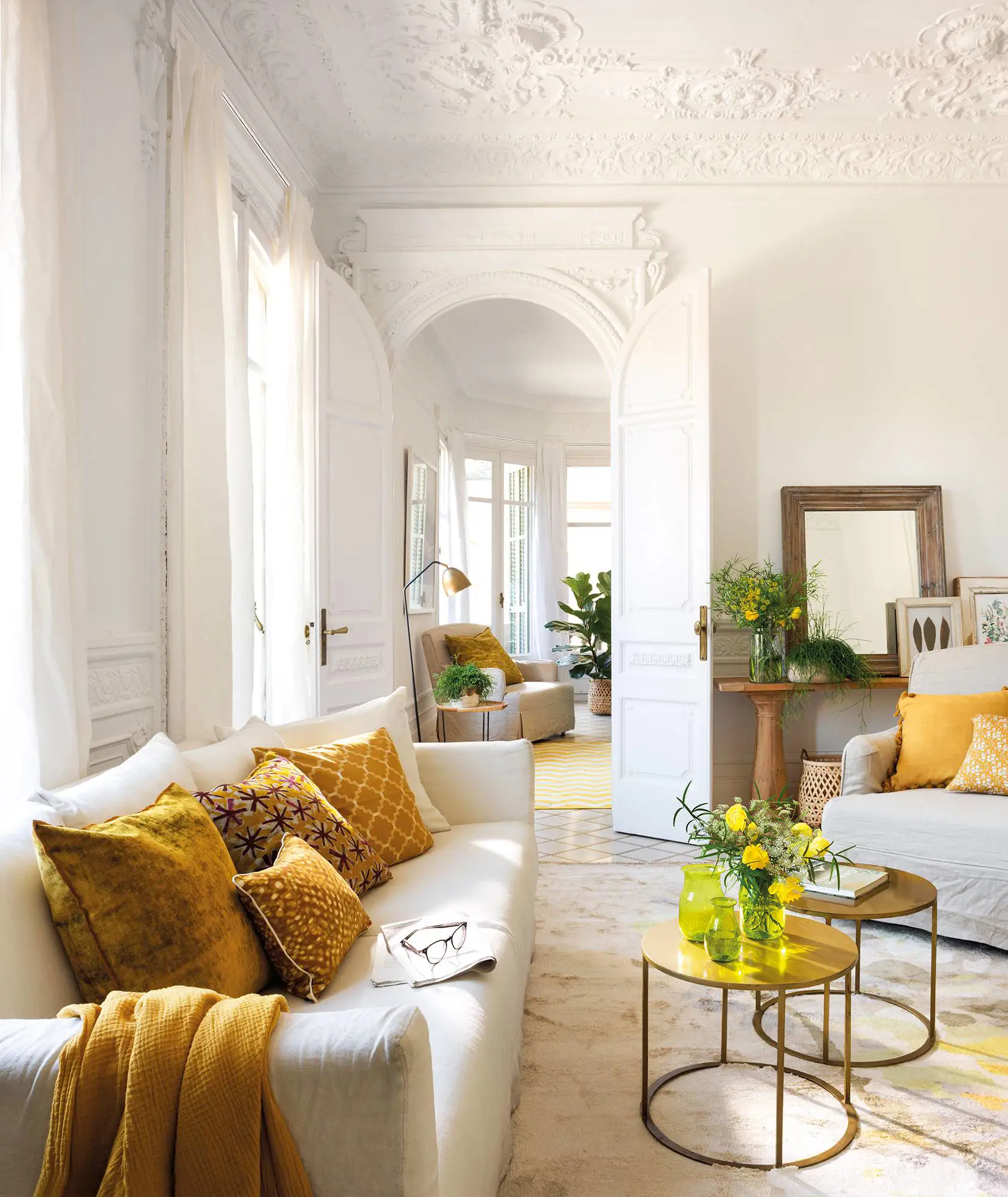 Salón clásico con sofá blanco, cojines amarillos y mesas doradas.