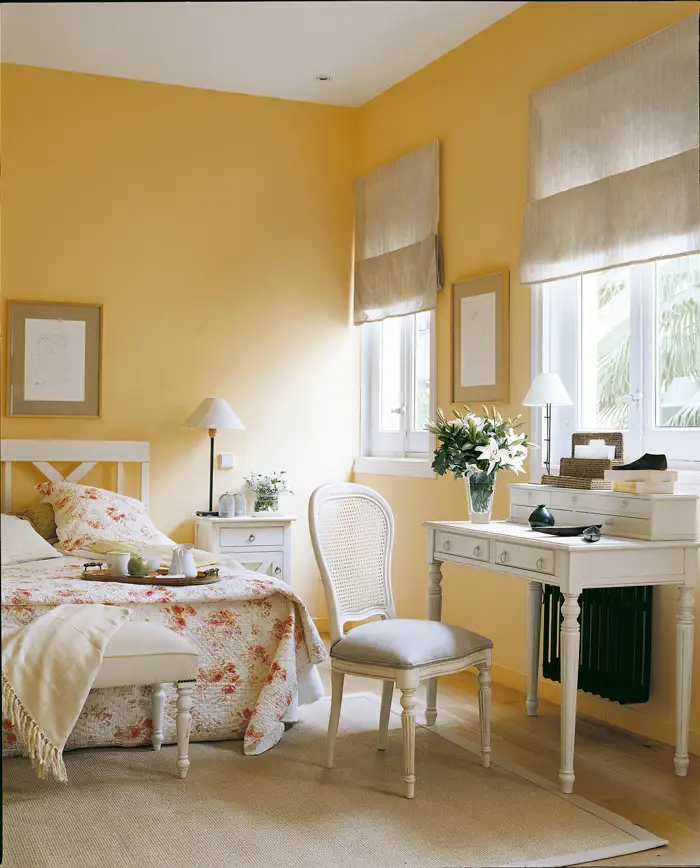 Dormitorio con paredes de color amarillo, colcha de flores, escritorio romántico blanco y silla con respaldo rejlla de mimbre