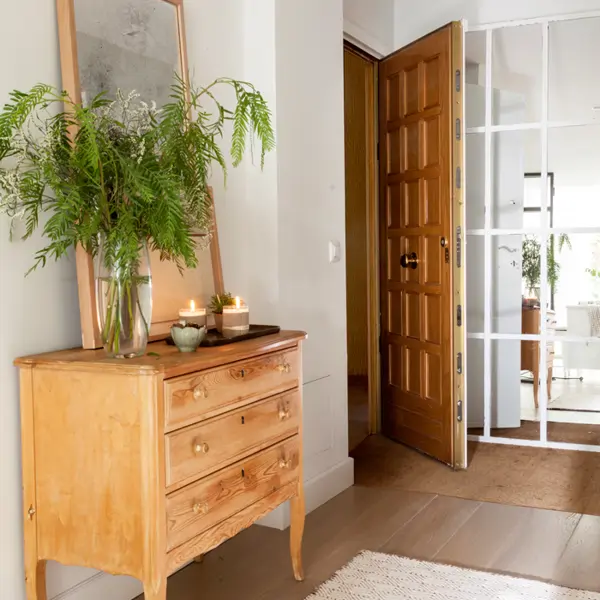 Recibidor con puerta de entrada abierta, cómoda de madera, espejo con marco de madera, velas encendidas y plantas_466563