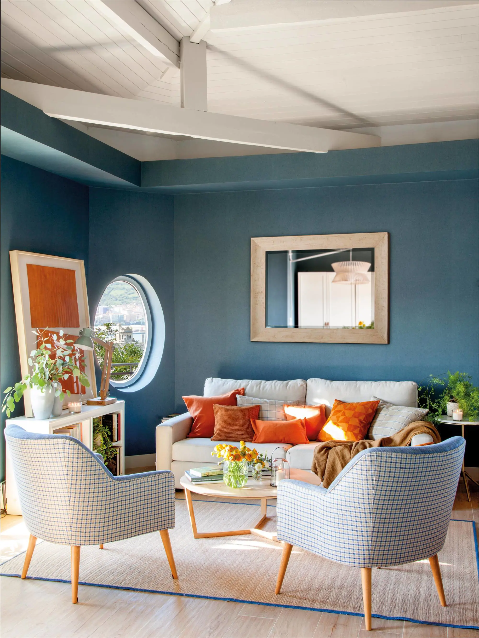 Salón en azul con vigas, sofá, butacas, espejo y ventana redonda
