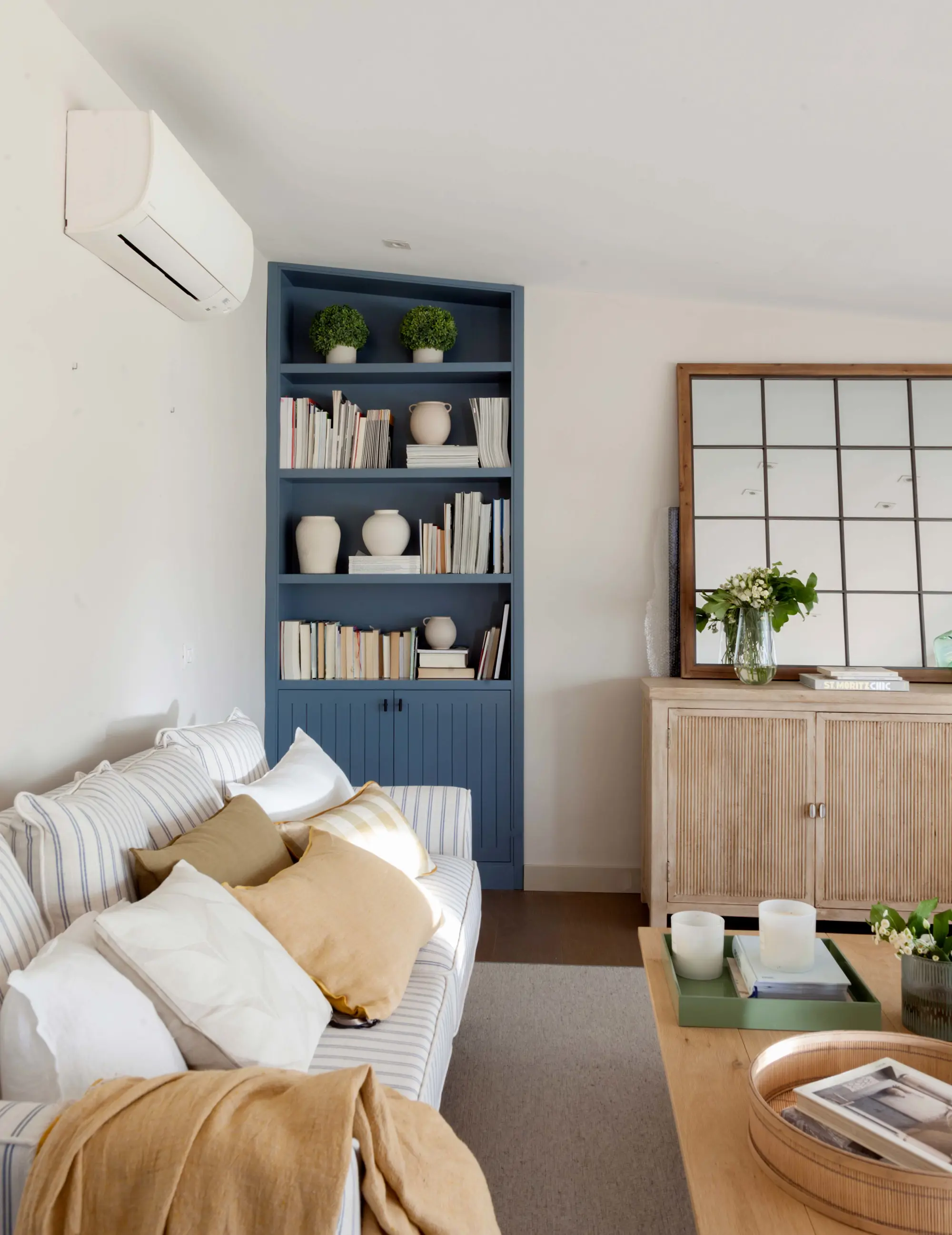 salón con sofá contra la pared, estantería azul, espejo de cuadrícula sobre aparador y aire acondicionado